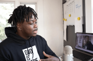 Man at home podcasting live with whiteboard in the background; Microphone and laptop in the foreground with camera on displaying his live stream-min