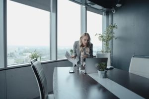 View stylish office corner with large windows with a woman leaning over conference table with empty chairs, talking into laptop