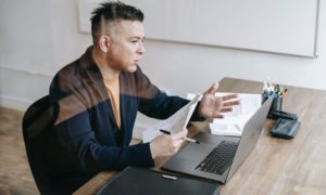 Side view of man seen through glass talking into laptop, holding a paper and pen with moving hands