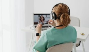 Back view of seated woman working at desk in front of laptop in white room beside curtained window, twiddling a pen and engaging with virtual teacher