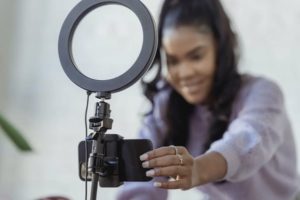 Blurry view of woman in background reaching with left arm to adjust device in tripod with ring light in foreground