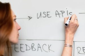 Close up view of woman’s focused face against whiteboard, scribbling down notes using a black marker in right hand, writing the words “Use APIs.