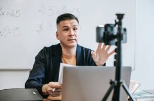 Man in front of laptop and interacting with mobile camera on tripod, teaching in front of whiteboard in classroom