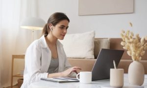 View of peaceful woman working on laptop, seated on floor in stylish beige colored living room at home