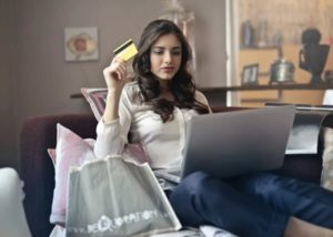 View of woman sitting comfortably on couch at home, surrounded by pillows and shopping bags, holding a credit card and shopping online from her laptop