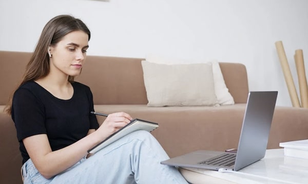 Calm young woman seated on floor against couch in serene and beige-colored room, writing in notebook in front of open laptop