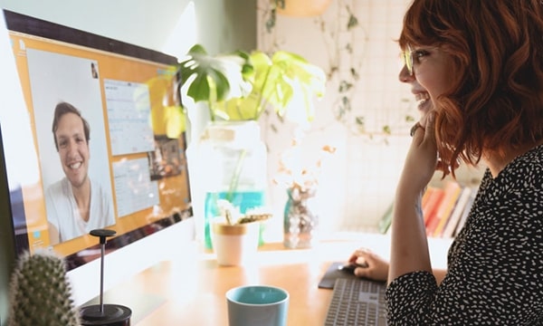 Side view of young woman laughing and working at desktop in sunny corner while video chatting a young man onscreen