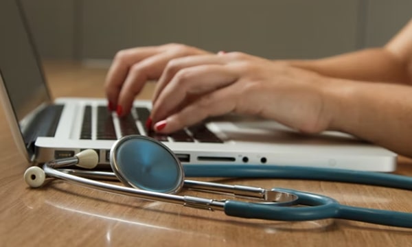 Side close-up view of hands typing away at open laptop on desk with stethoscope in foreground