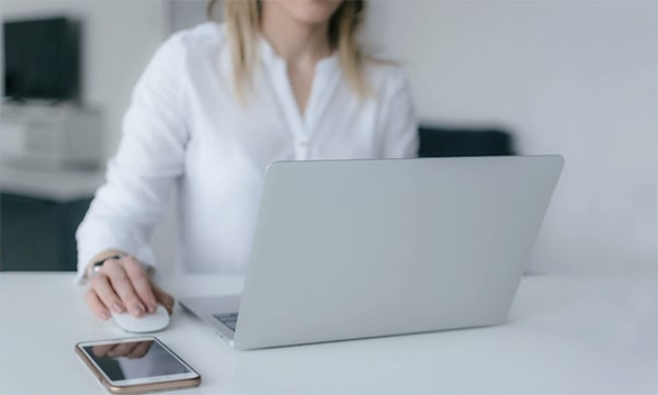 Business casual woman seated and working on laptop at a table with mouse and iPhone at table in a computer lab