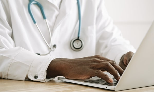 View of doctor in a lab coat with stethoscope, seated and typing on a laptop